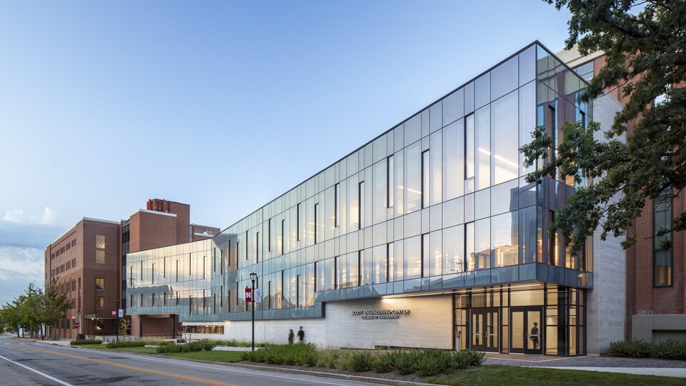 Modern glass building with reflective windows on a quiet street, trees, and a clear sky.