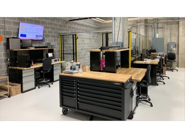 Modern industrial workspace with desks, chairs, storage cabinets, and machinery in a concrete-walled room.